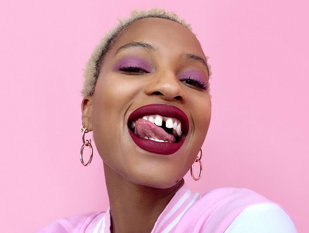Close-up of a Black model with pink eyeshadow and berry lips smiling and licking her teeth against pink background Desktop