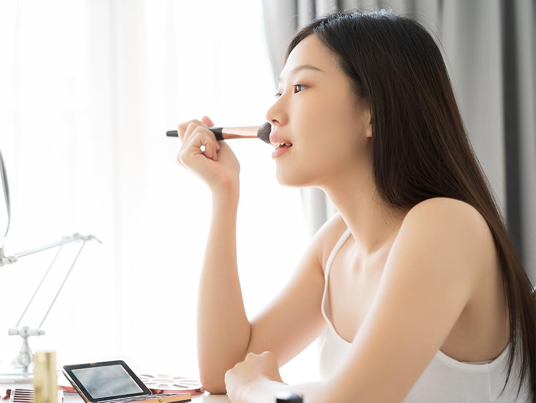 A photo of a young woman applying makeup with a brush on her cheek wearing a white spaghetti top Desktop