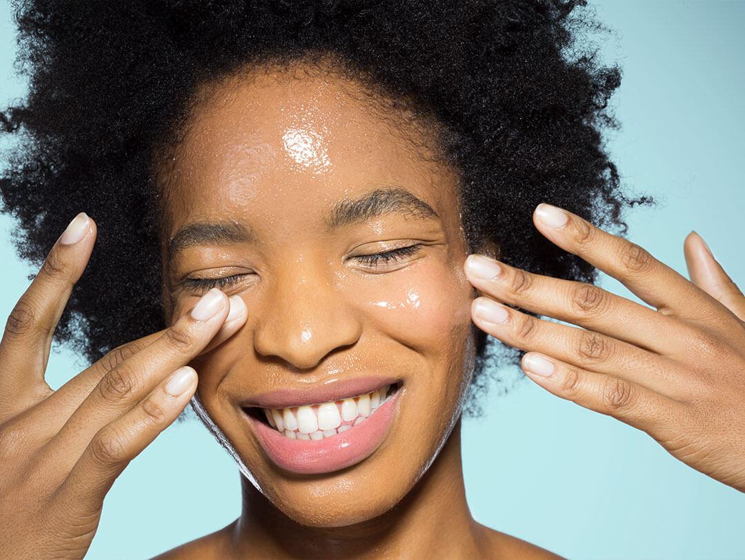 A close-up image of a woman with color and afro hair smiling directly at the camera while applying moisturizer Desktop