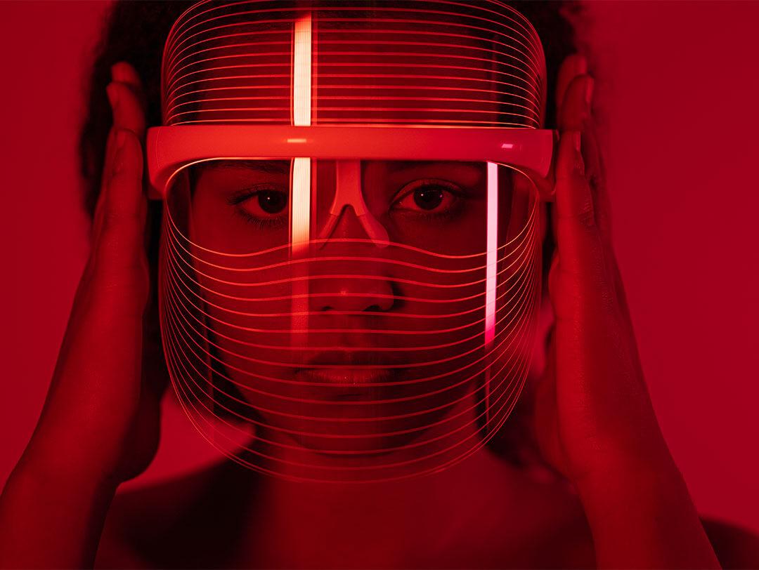 A close-up image of a woman wearing a red-light therapy mask while situated in a room illuminated with red lighting Desktop