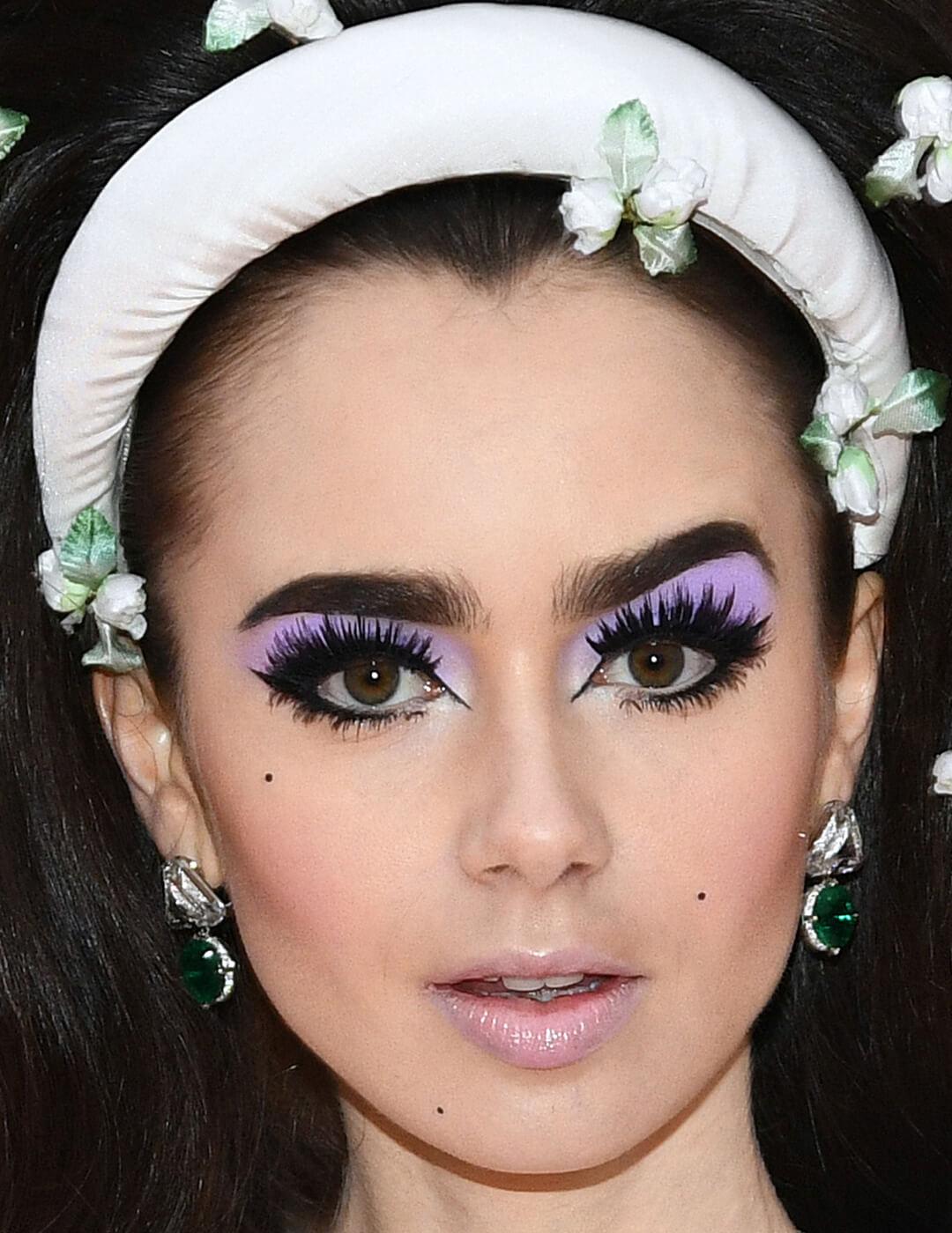 Lily Collins looking retro in a purple eyeshadow makeup look, glossy and frosty nude lips, and white headband with flowers Mobile
