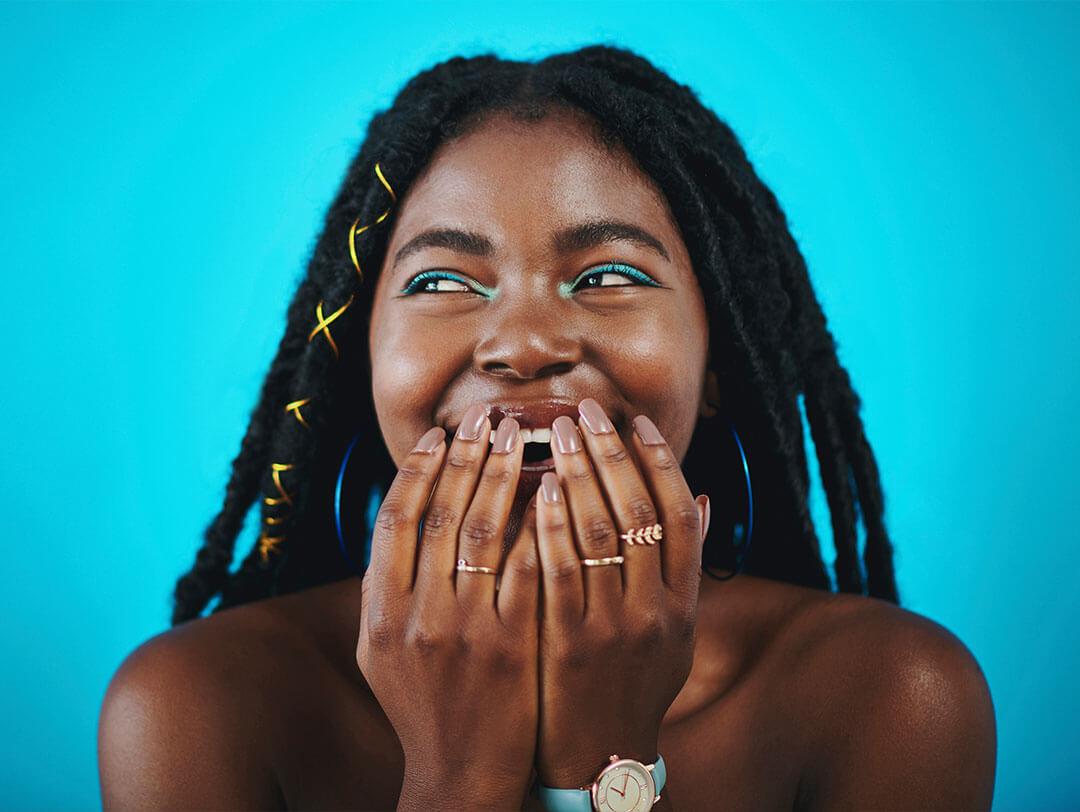 Woman of color with blue eyeshadow, hands almost covering her mouth, giggling against a blue backdrop Desktop