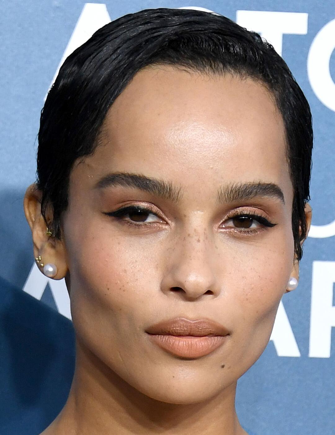 A closeup of Zoë Kravitz with pixie cut hairstyle, wearing black eyeliner and shimmery brown eyeshadow with a nude lipstick color Mobile