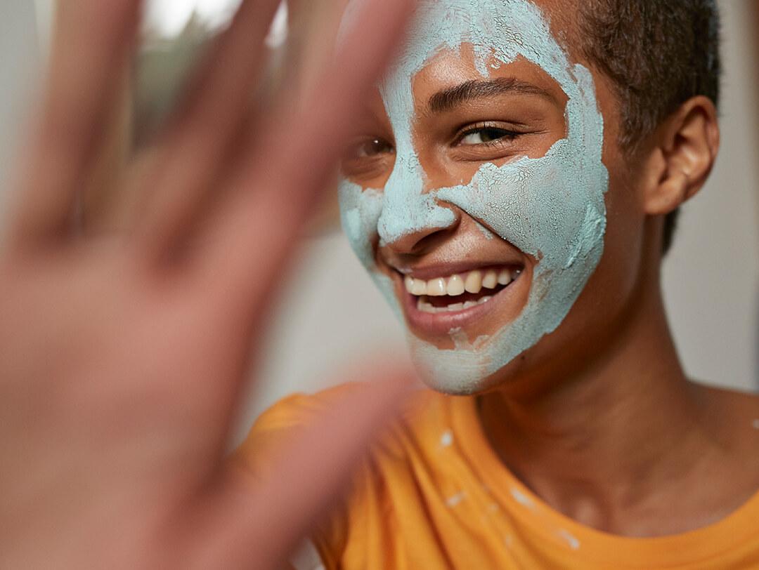 A focused photo of a woman with a teal mud mask on her face and a blurry photo of her hand Desktop