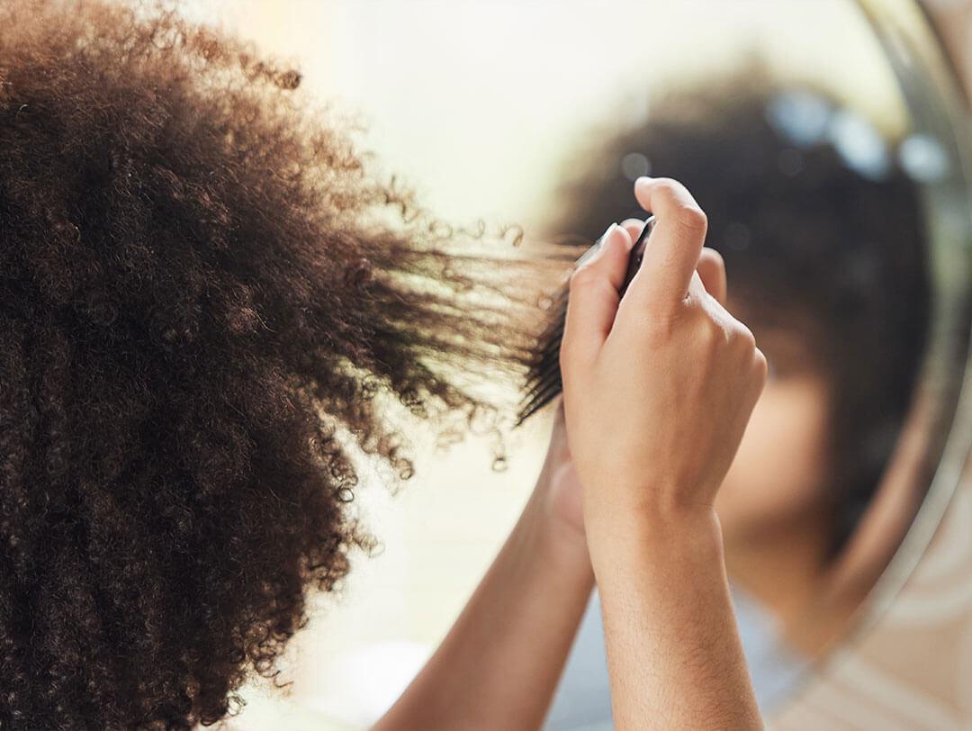 A person using their hands and a detangling tool to carefully work through thick, curly hair in front of a mirror, showing the effort to treat and prevent single-strand fairy knots Desktop