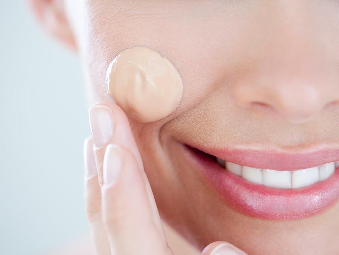 Close-up image of a smiling woman applying liquid foundation on her cheek Desktop