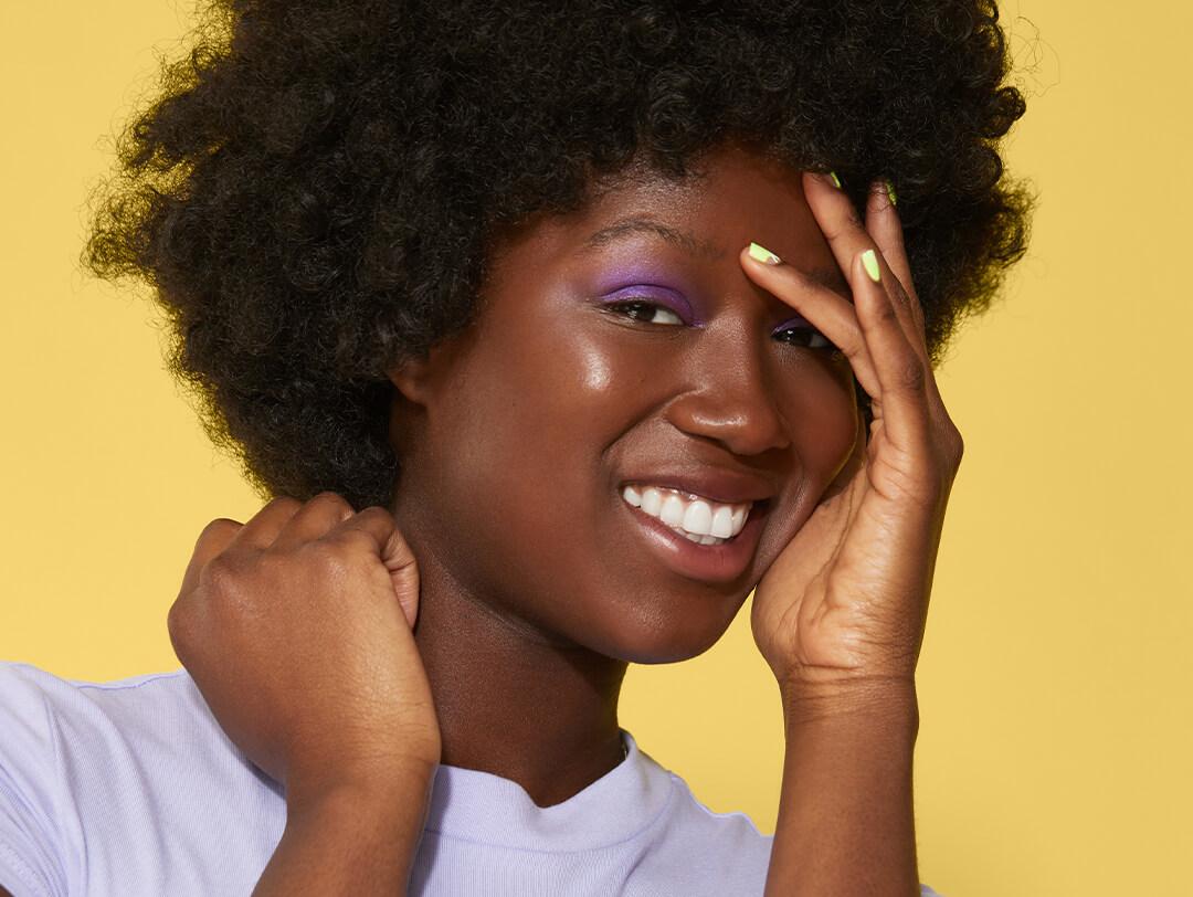 Black model rocking a purple eyeshadow look and lime green nails smiling and covering one eye with her hand Desktop