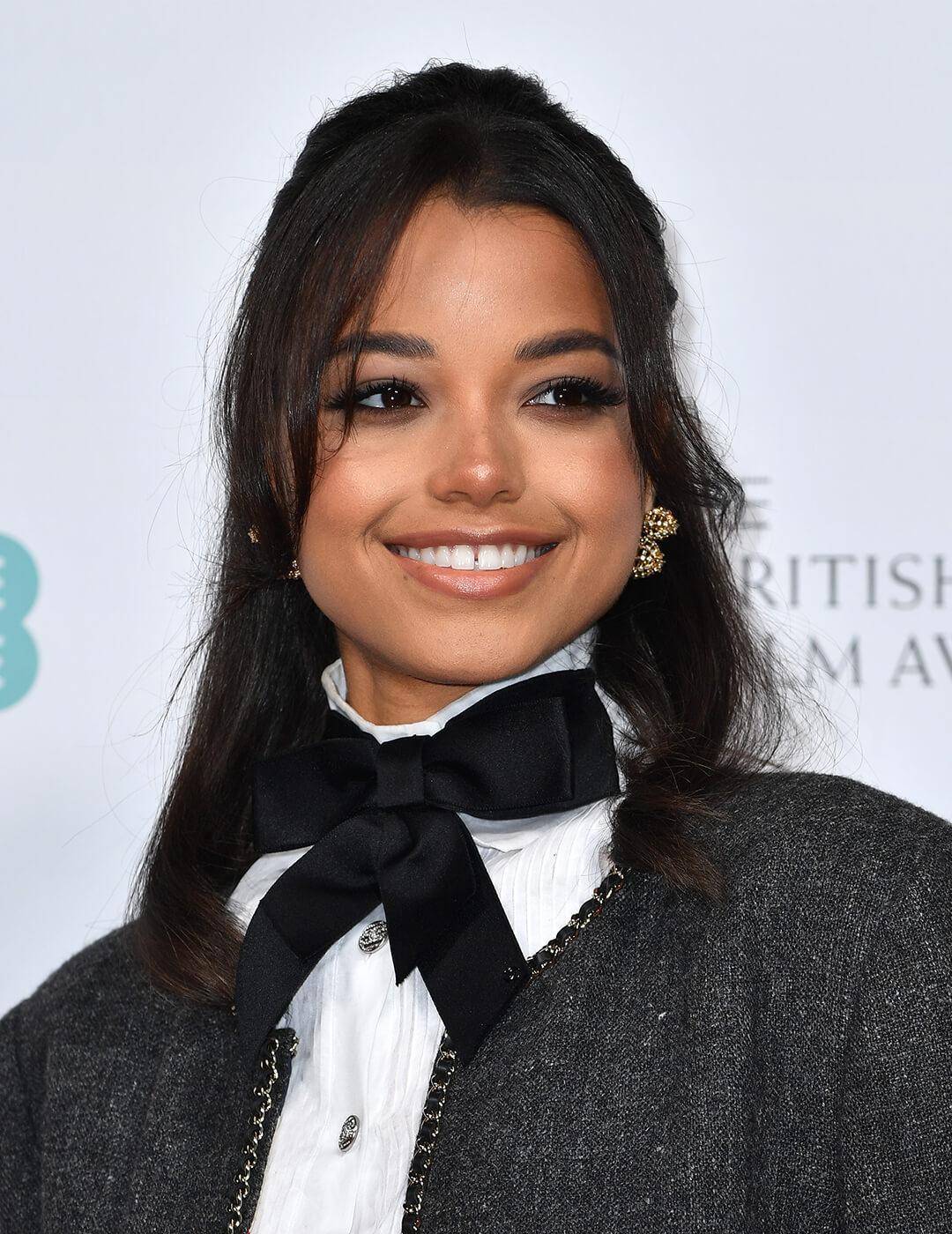 A photo of Ella Balinska wearing a bright smile in her nude lipstick, black bow tie, gold earrings with her black hair pulling a half-up, half-down hairstyle Mobile