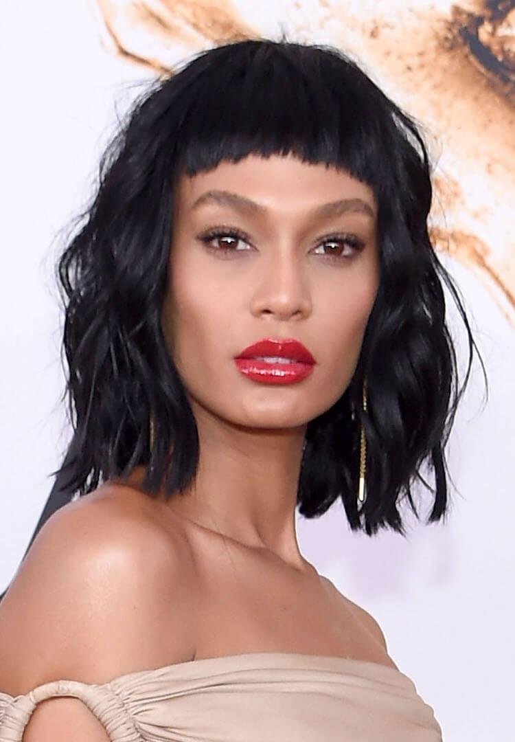A photo of model Joan Smalls donning a chic nude off-shoulder dress with her striking black micro bangs wearing a bold swipe of vibrant red lipstick adorned with stylish bangles Desktop