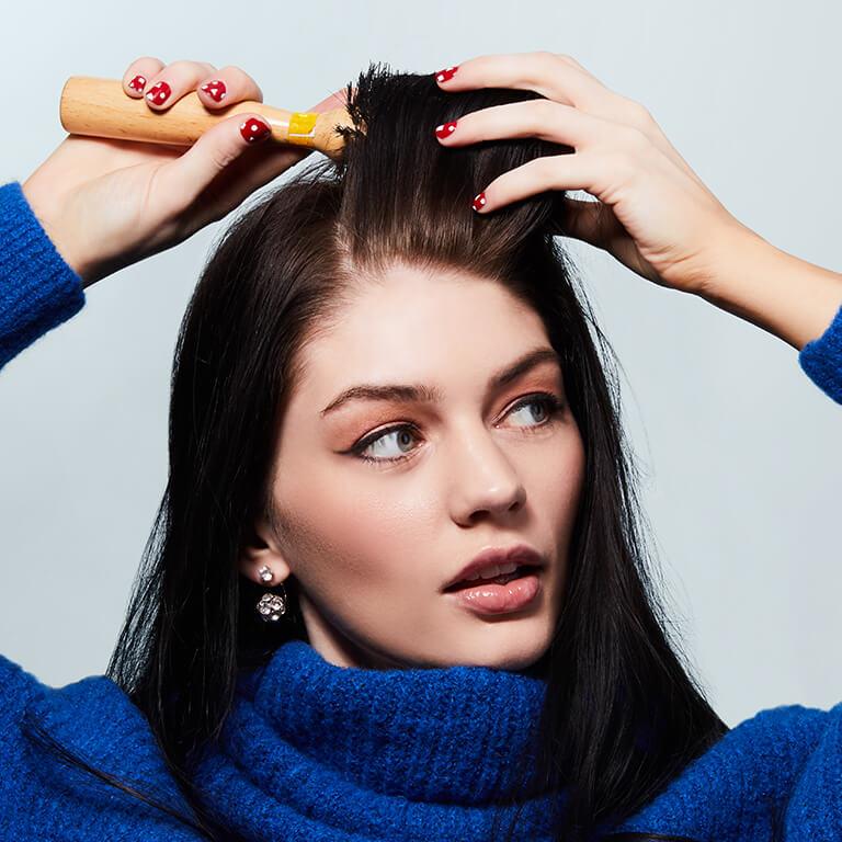 A closeup image of a model wearing blue knitted top brushing her hair with a round brush Mobile