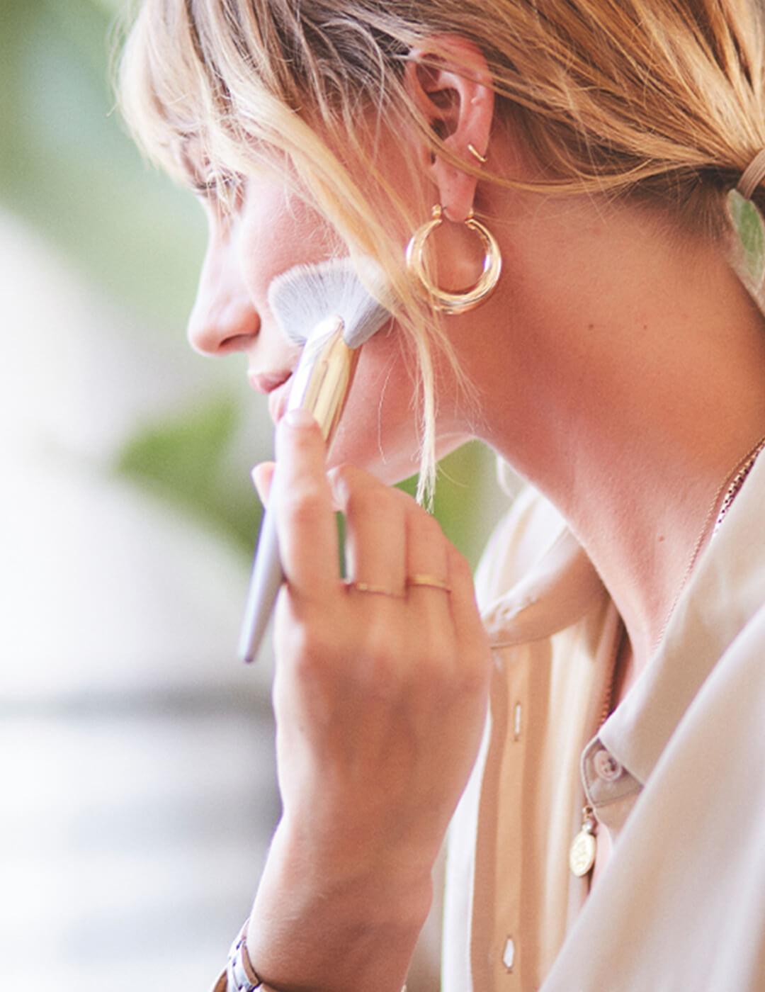 An image of a blond woman facing sideways holding a highlighter brush with gold earrings Mobile