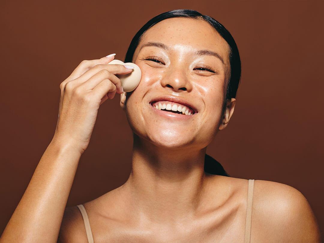 A picture of a happy woman applying a mixture of foundation and moisturizer with a beauty sponge to her face Desktop