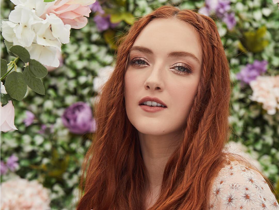 Close-up image of a model with copper hair in a floral outfit and wearing rose gold eyeshadow makeup against a garden background Desktop