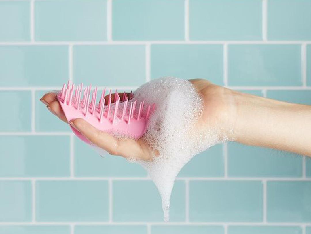 Woman's hand holding a pink scalp massager against teal bathroom tiles Desktop