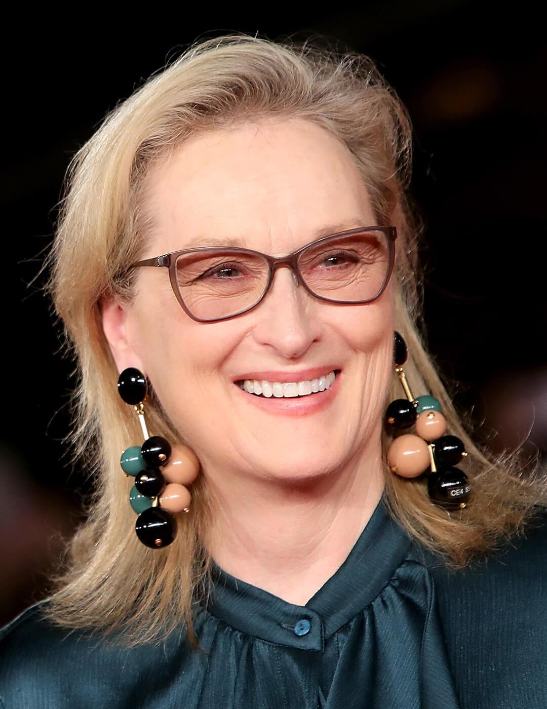 A photo of Meryl Streep wearing brown glasses, ball-drop earrings and a dark green blouse Mobile