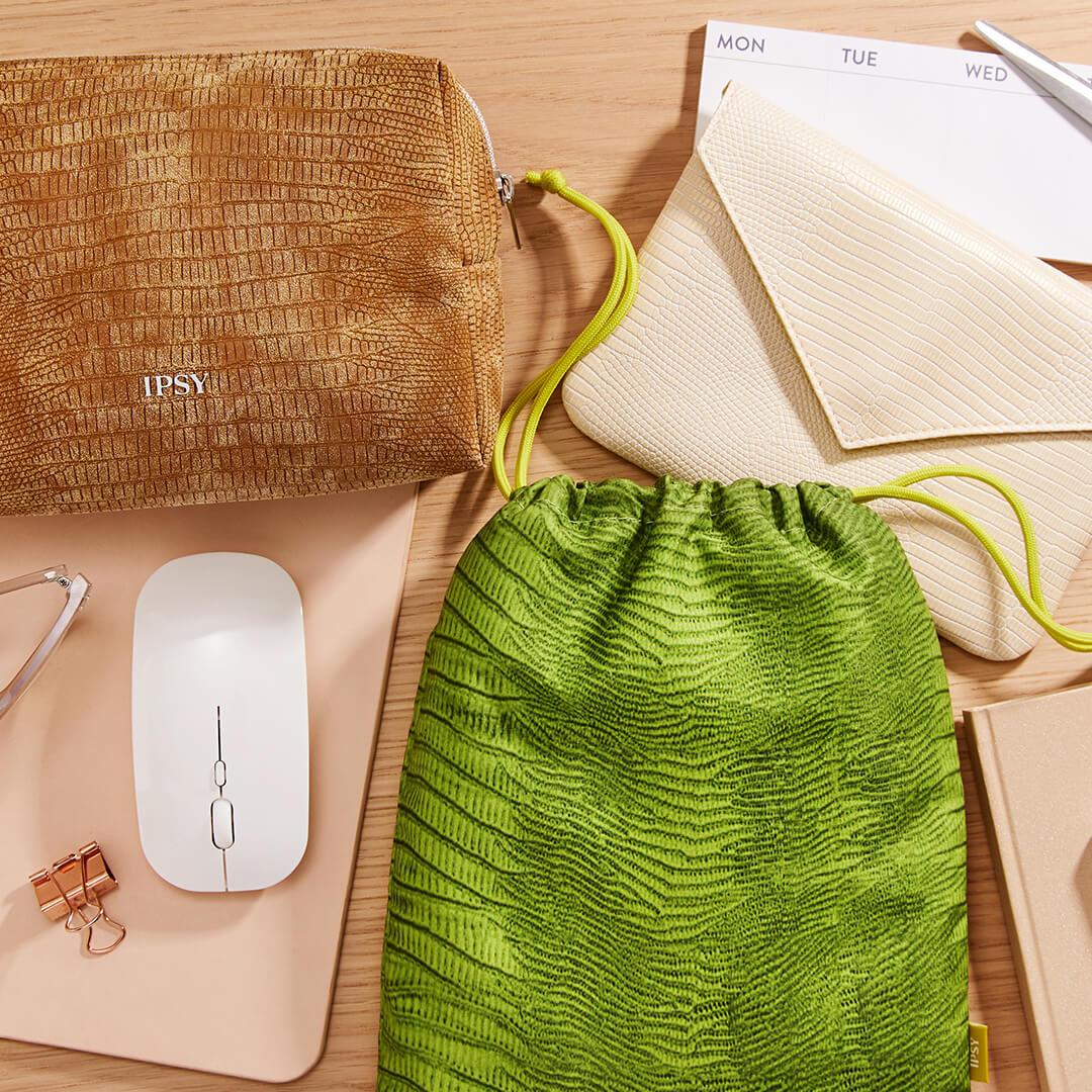 Flat-lay image of the August 2021 IPSY Glam Bag, Glam Bag Plus, and Glam Bag X bags together with a wireless mouse, paper clip, notebooks, and calendar Mobile