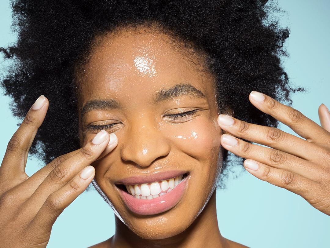 Close-up of young mixed-race woman applying glossy face make-up moisturizer, smiling, with eyes closed with turquoise background Desktop