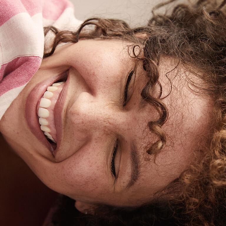 A closeup image of a model with curly hair smiling big and a closeup image of a model with curly hair wearing a lavender eye makeup look Mobile