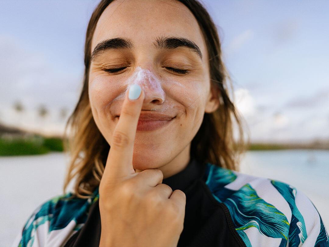 A woman is shown in a picture applying sunscreen to her nose, making amusing facial expressions with her eyes closed, while being photographed by a camera Desktop