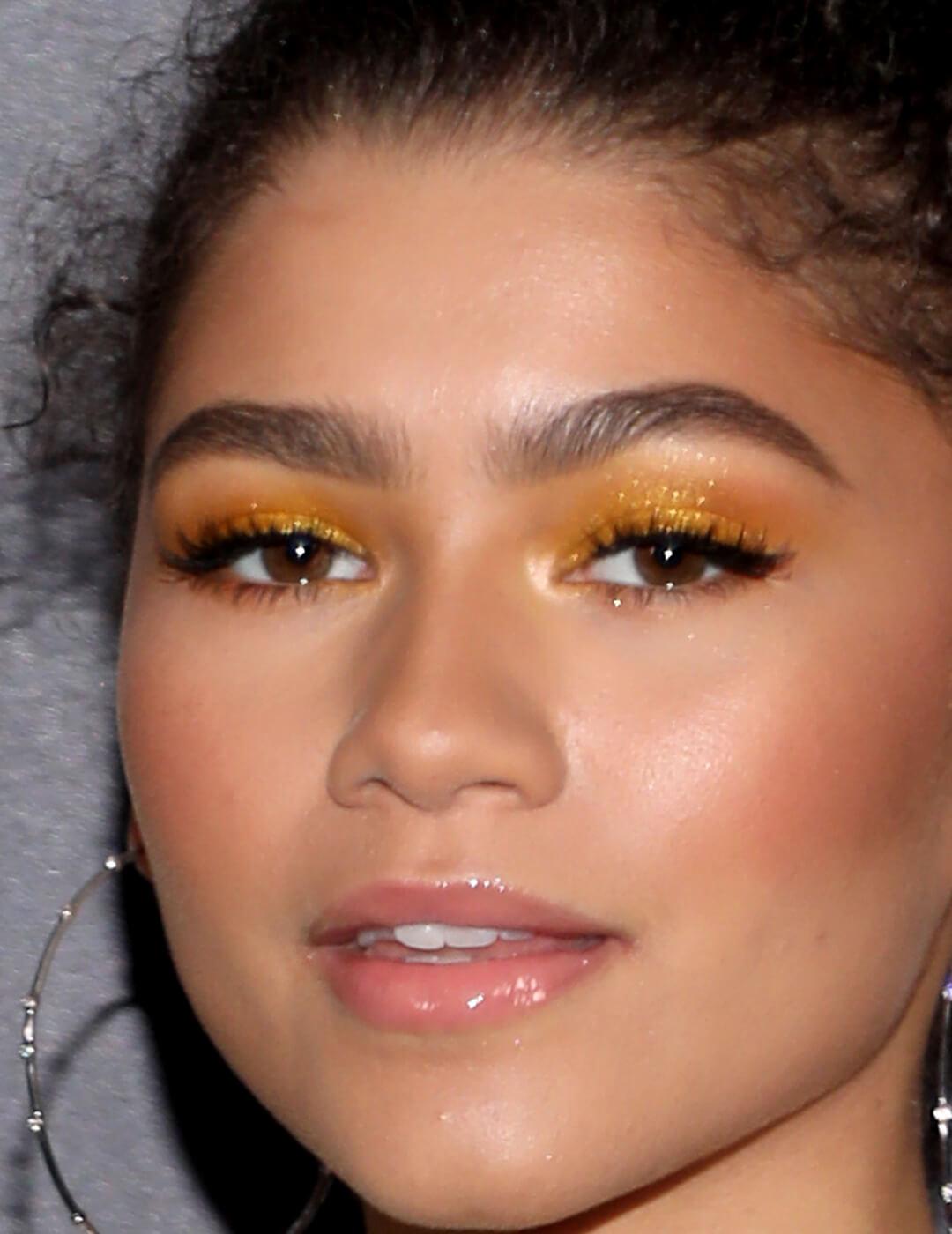 Close-up image of Zendaya wearing a shimmery gold-yellow eye makeup look paired with nude lips and big hoop earrings Desktop