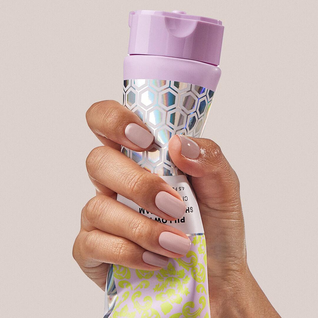 A closeup photo of a hand squeezing a tube of REFRESHMENTS Pillowy Shave Cream on a beige background Desktop