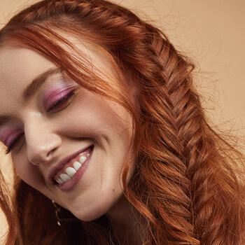 A close-up image of a red-haired model rocking pink eyeshadow look and messy fishtail braids Mobile