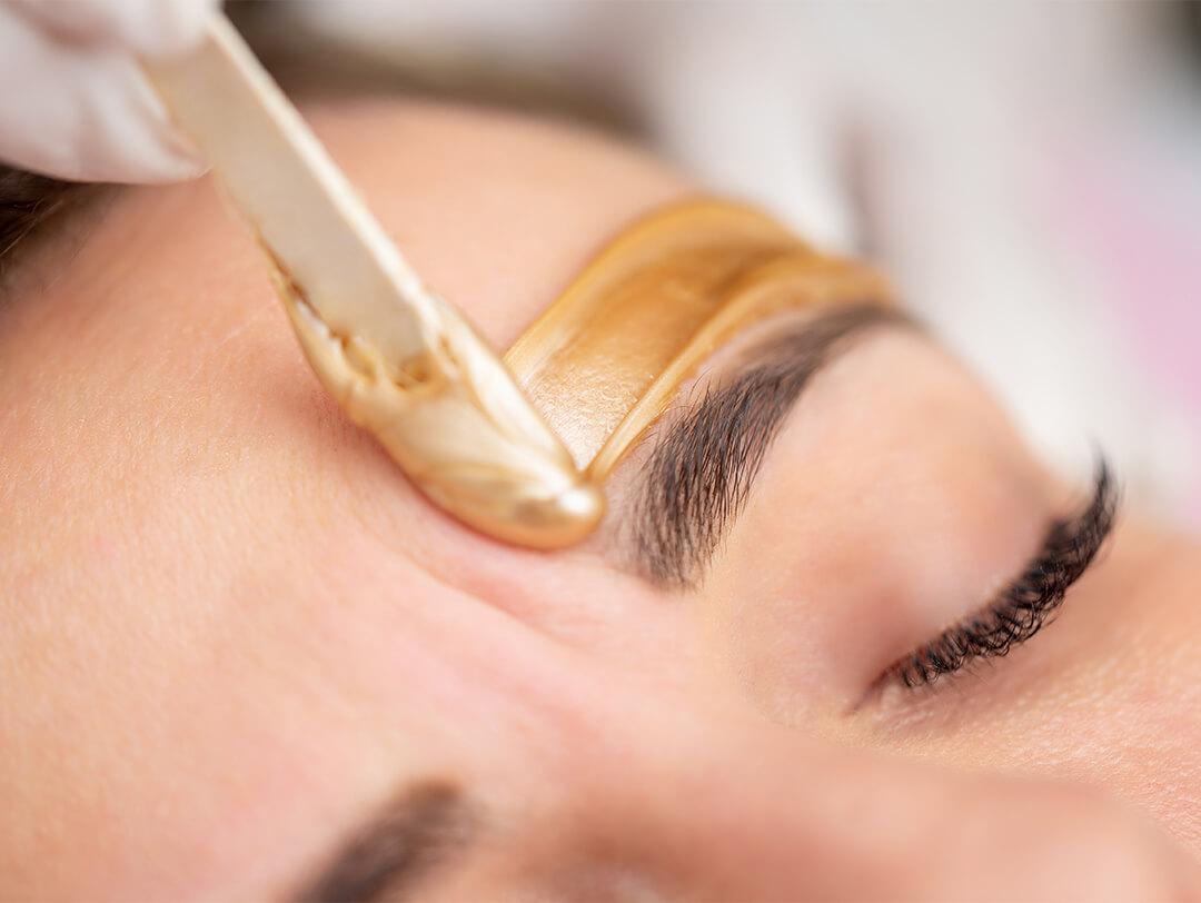 Image of gold wax applied with a spatula above woman's eyebrow Desktop