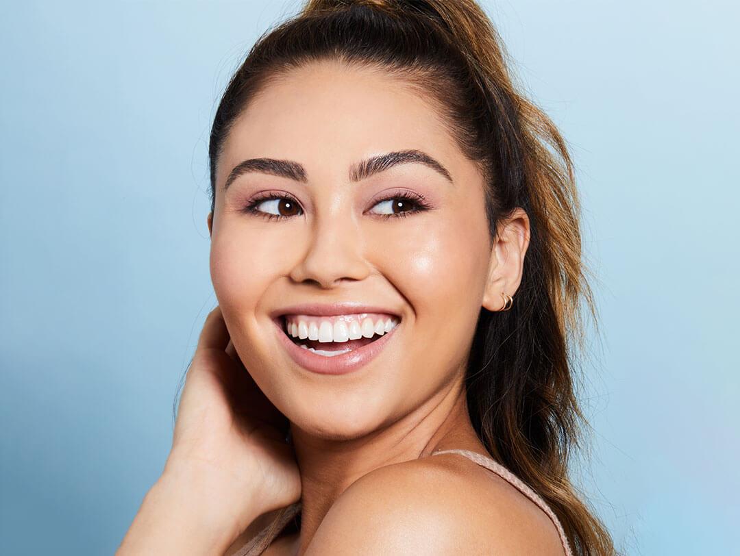 An image of a woman with a sideways gaze, displaying a cheerful smile directed at the camera, wearing a lightweight foundation and makeup, completing her look with golden round earrings Desktop