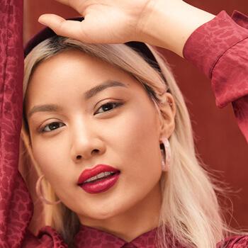 An image of a model wearing pink hoop earrings, red lipstick, and black headband is raising her hand Mobile
