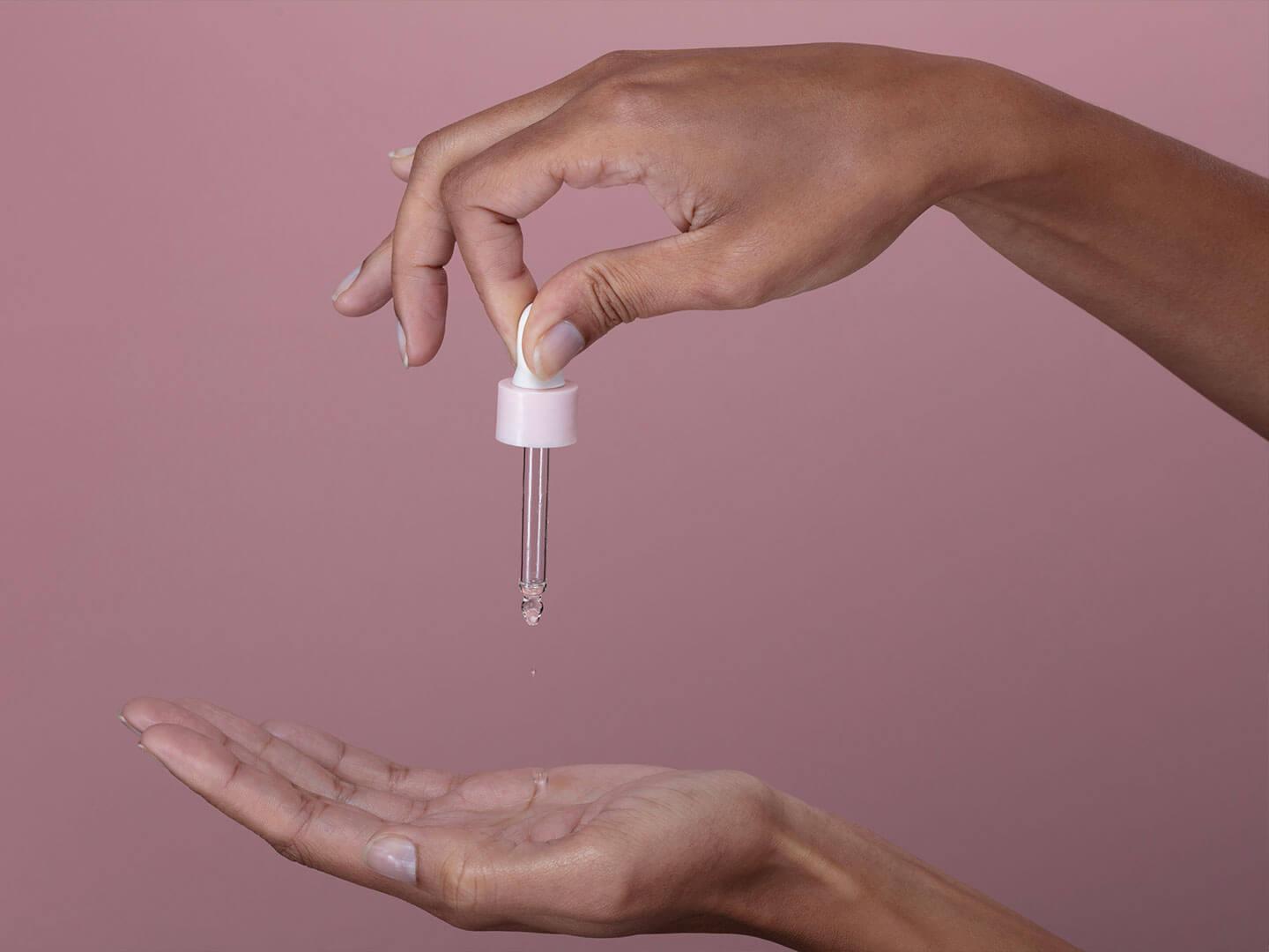 A close-up of hands delicately holding a glass dropper, with a few drops of a clear liquid falling into the palm against a soft pink background Desktop
