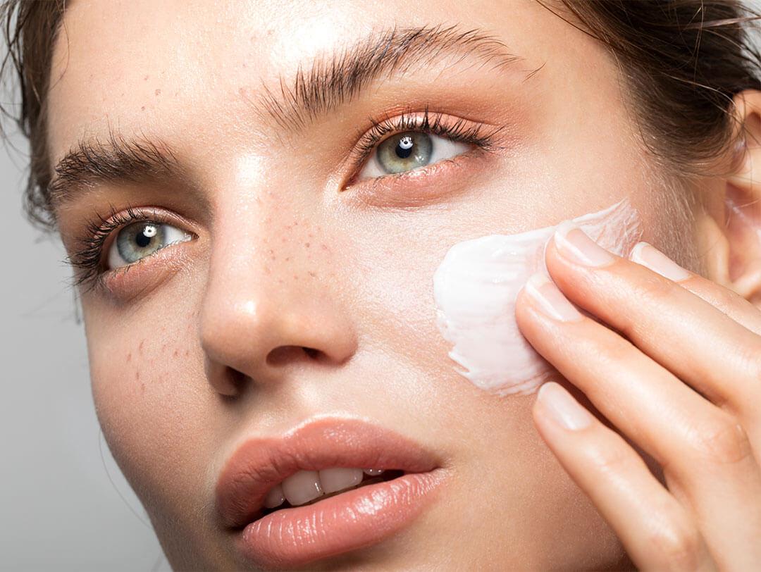 An up-close photo of a young woman applying cream on her face who has clear skin and a hint of freckles Desktop