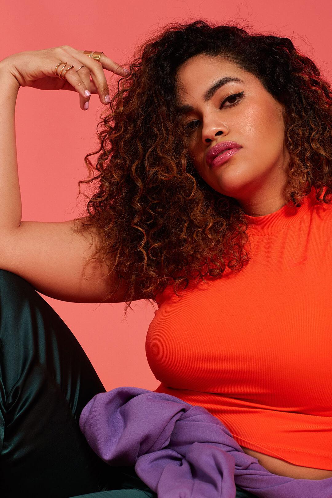 A photo of a woman with curly hair wearing an orange top on a pink background Desktop