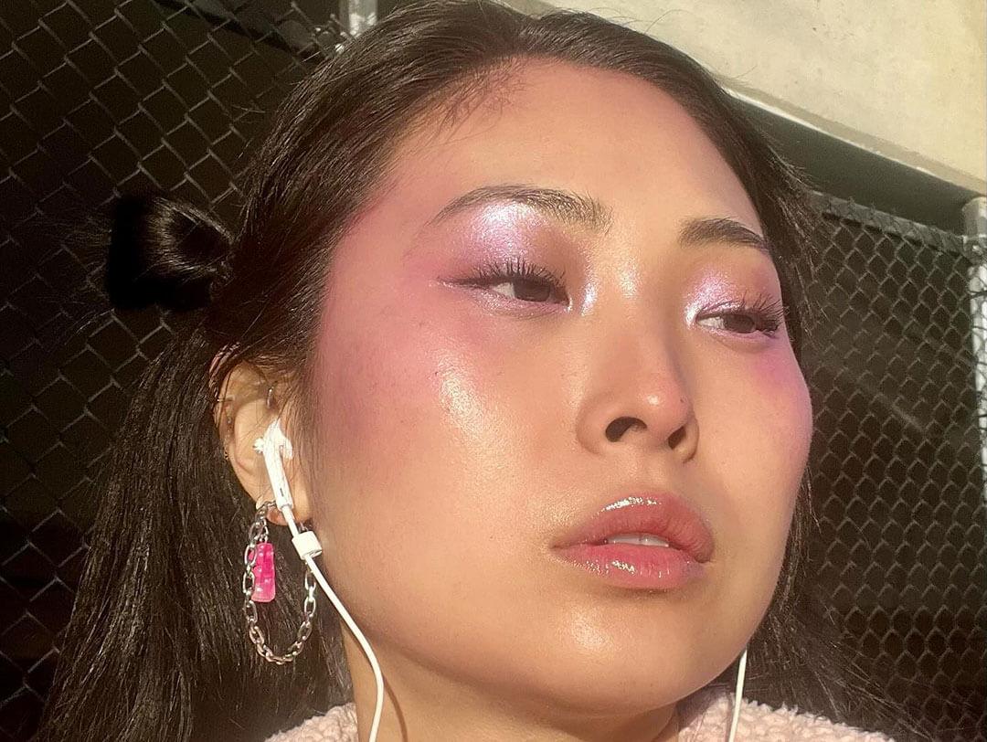 A woman with a radiant complexion, adorned with purple blush and shimmering eye makeup, glossy lips, and a subtle, dewy finish on her skin, illuminated by natural sunlight Desktop