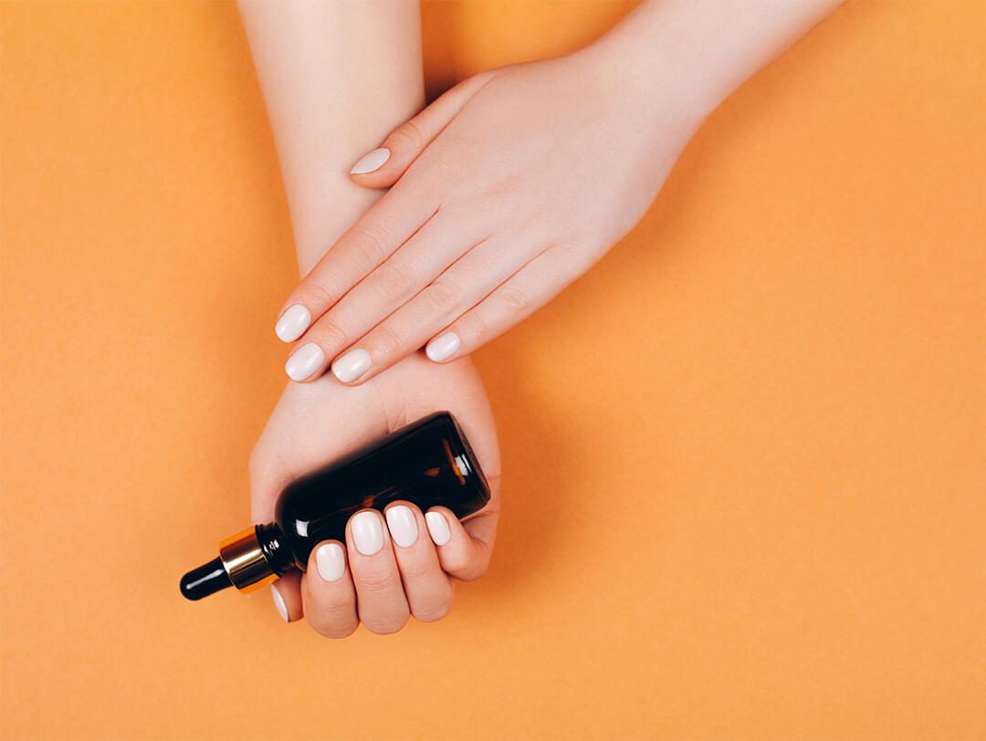 Image of two hands with white nail polish and one is holding a black bottle with dropper on orange background Desktop