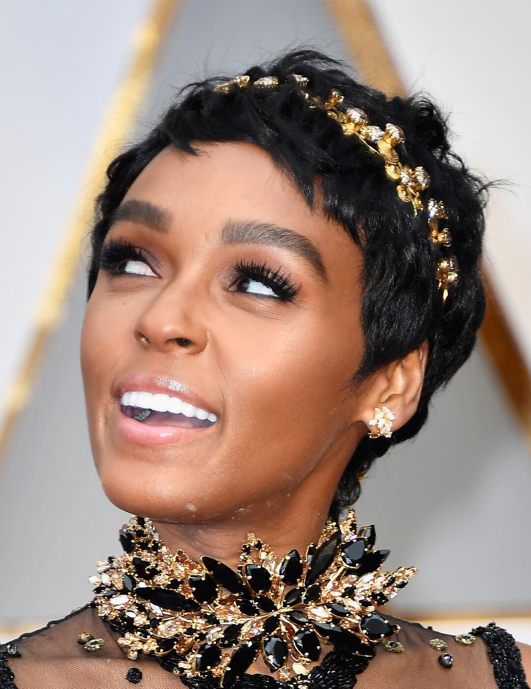 Close-up of Janelle Monae smiling while rocking a neutral makeup look, pixie cut hairstyle accented with gold and silver headband, and black and gold choker necklace Mobile