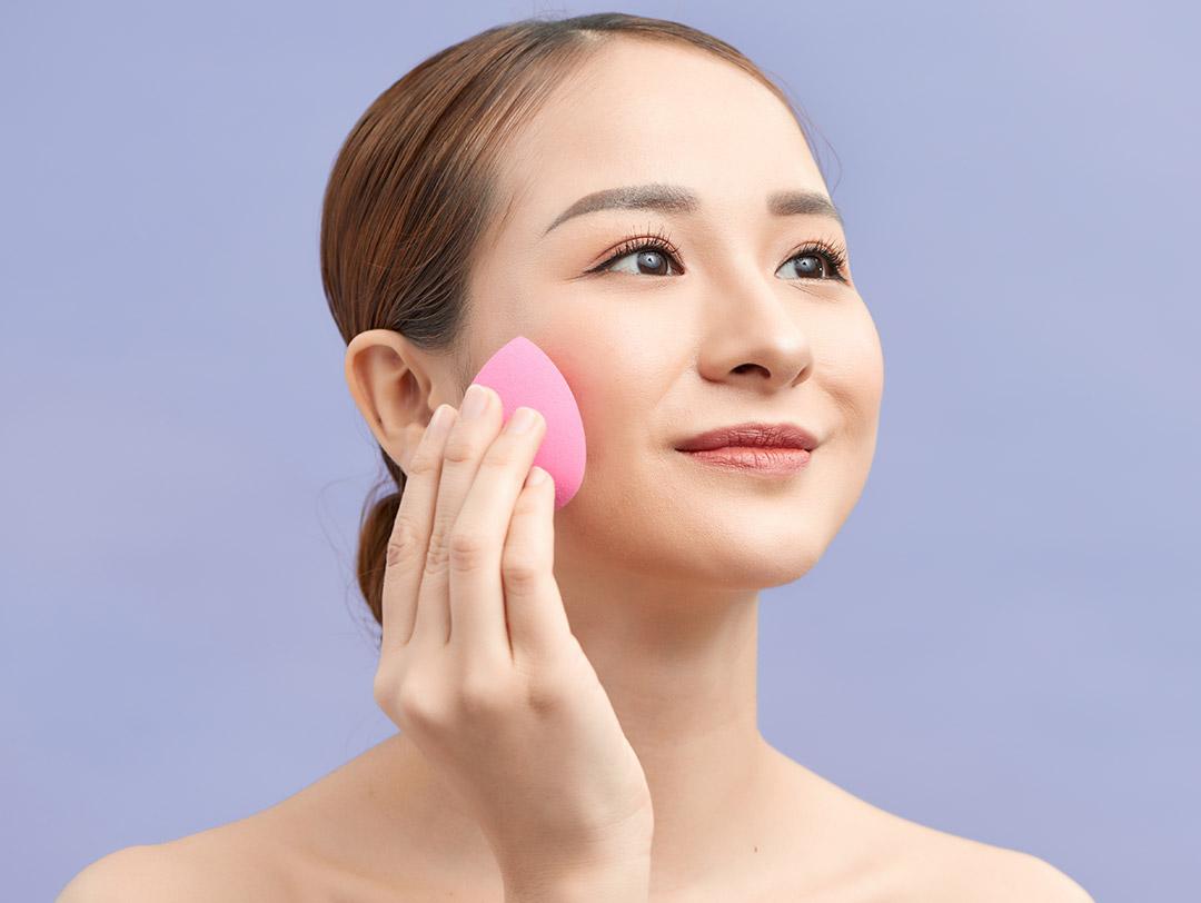 Close-up of a beautiful Asian woman putting on face makeup using a beauty sponge on lilac background Desktop