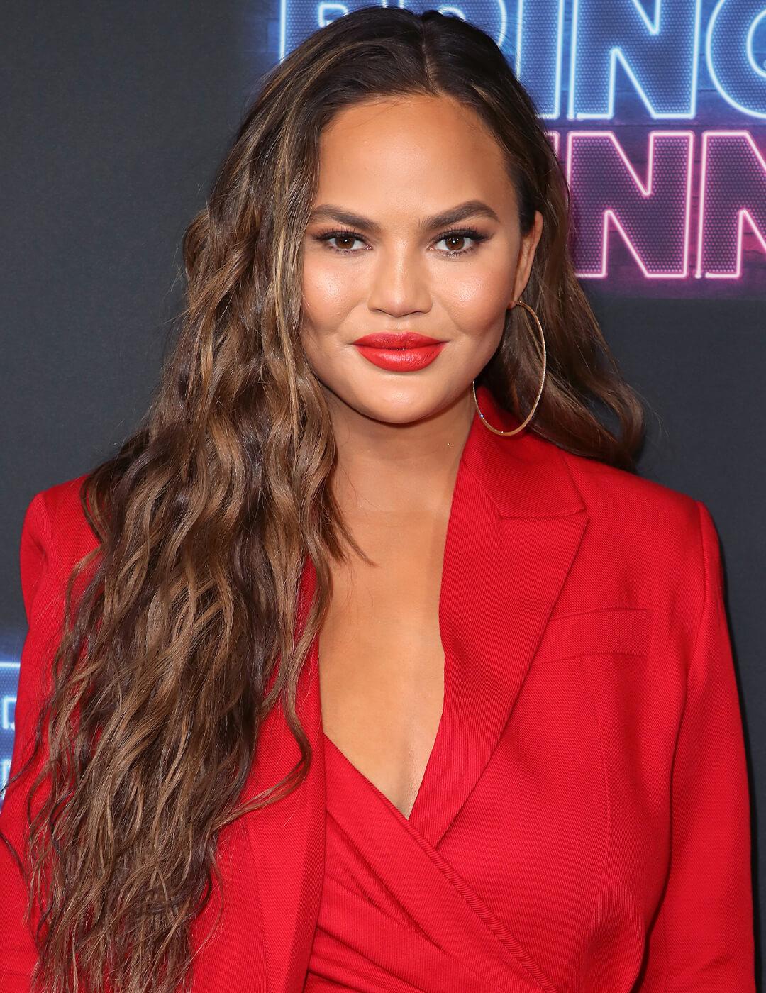 Chrissy Teigen rocking a long and wavy hairstyle with bold red lips and big hoop earrings in a red suit Mobile