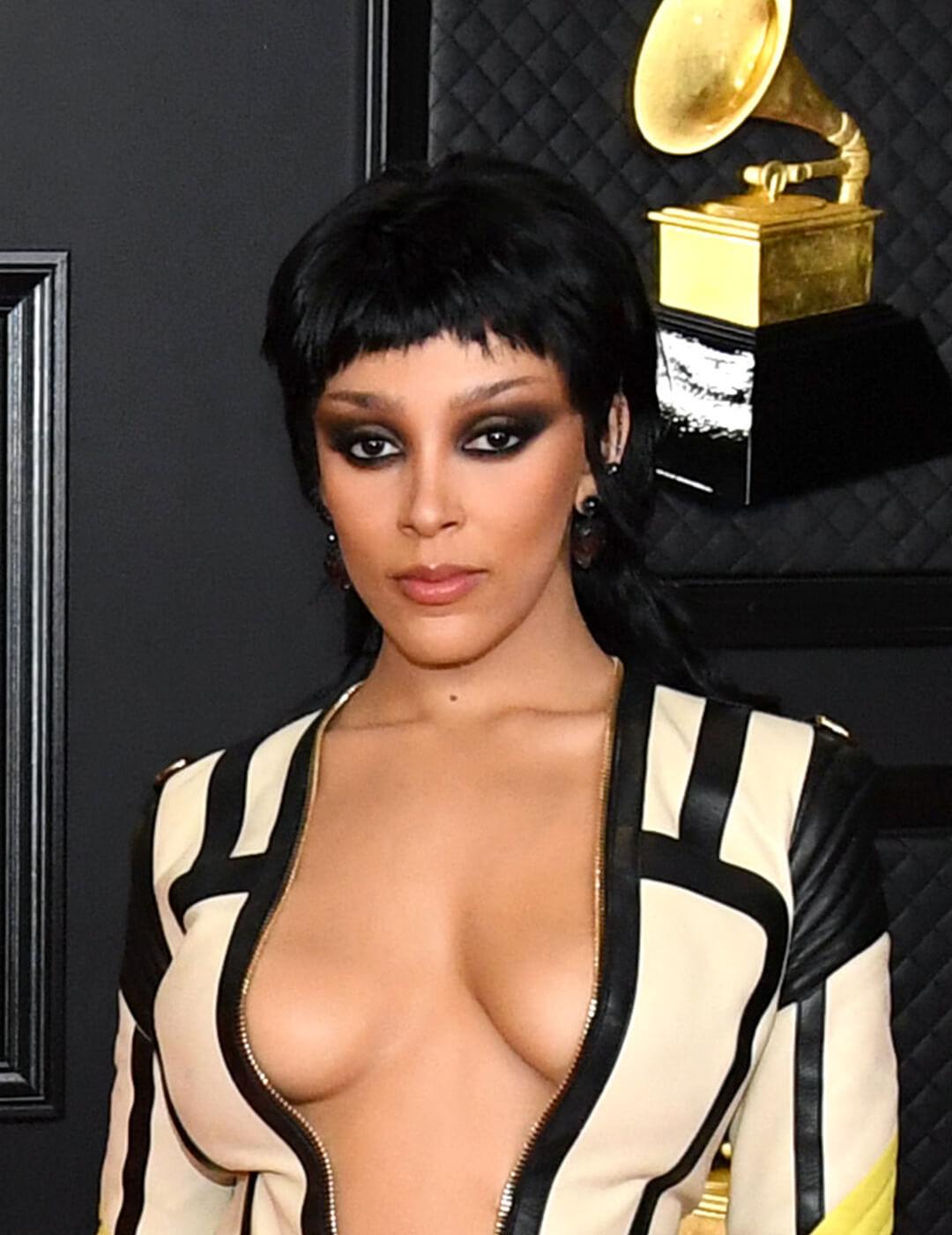 A photo of Doja Cat wearing a long sleeve zippered dress with a neon feathers and a plunging neckline along with her slicked-back, rocker-chic mullet cut Mobile