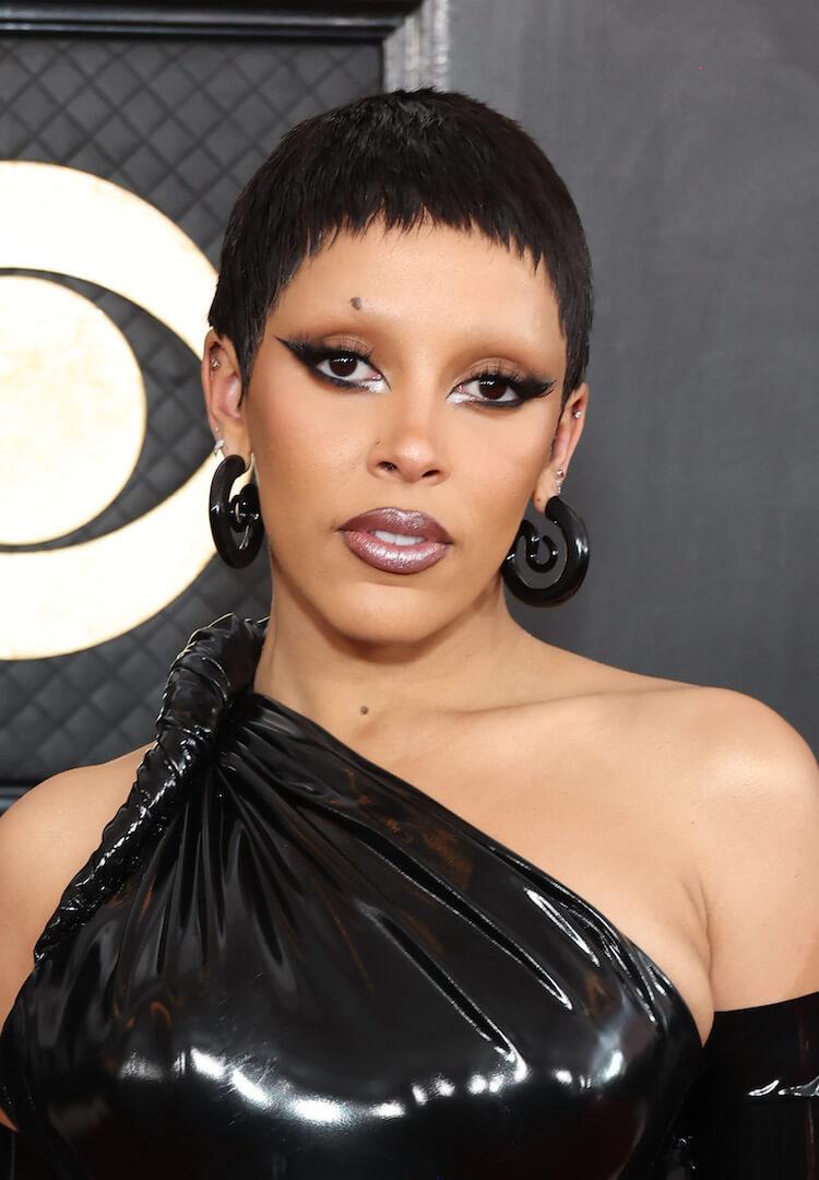 A photo Doja Cat rocks a mullet-style bangs hairstyle dressed in a sleek leather one-shoulder long black dress with a striking black liner paired with a unique black spiral-shaped earring Desktop