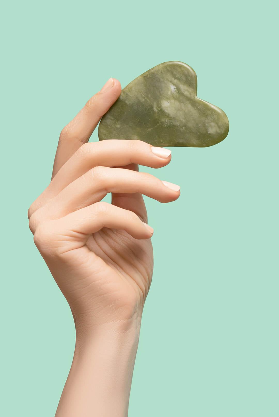 A photo of a hand holding a green jade gua sha on a green background Desktop