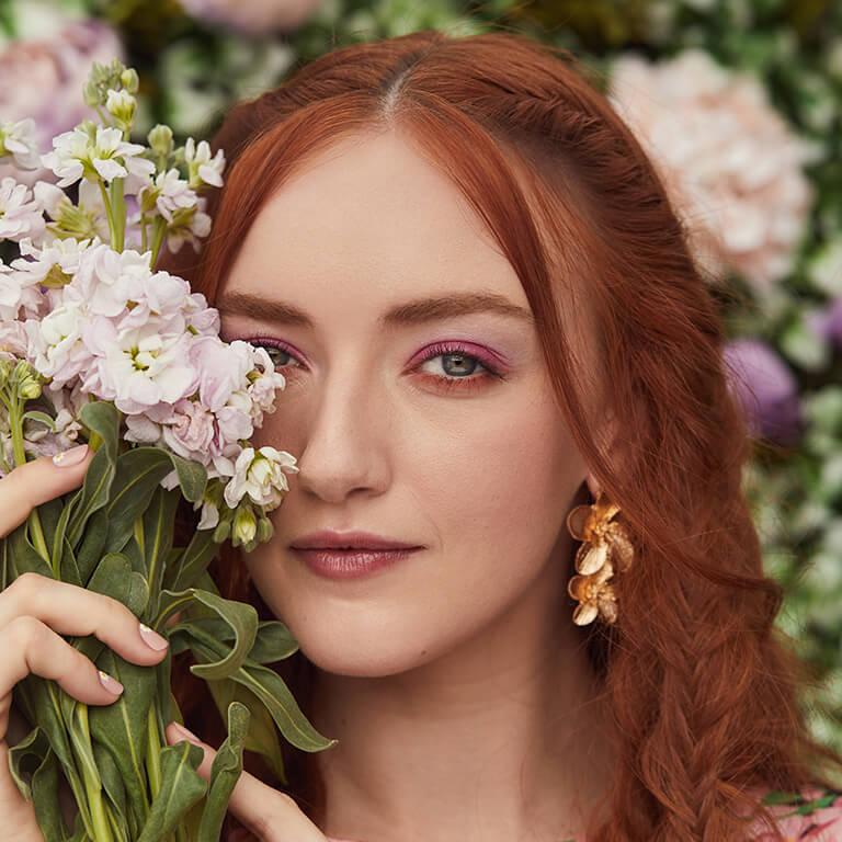 A close-up image of a red-haired model rocking a matte fuschia and peach eyeshadow look and floral earrings while covering half of her face with a bouquet of flowers Desktop