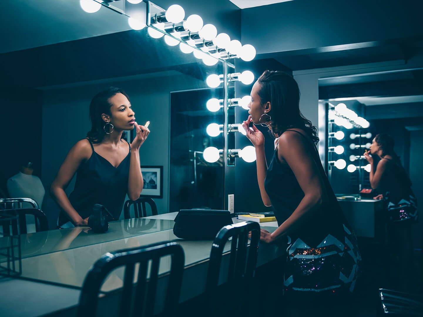 An image of a woman of color applying lipstick in front of a brightly lit vanity mirror, dressed in a black satin tank top and sporting a hoop earring Desktop