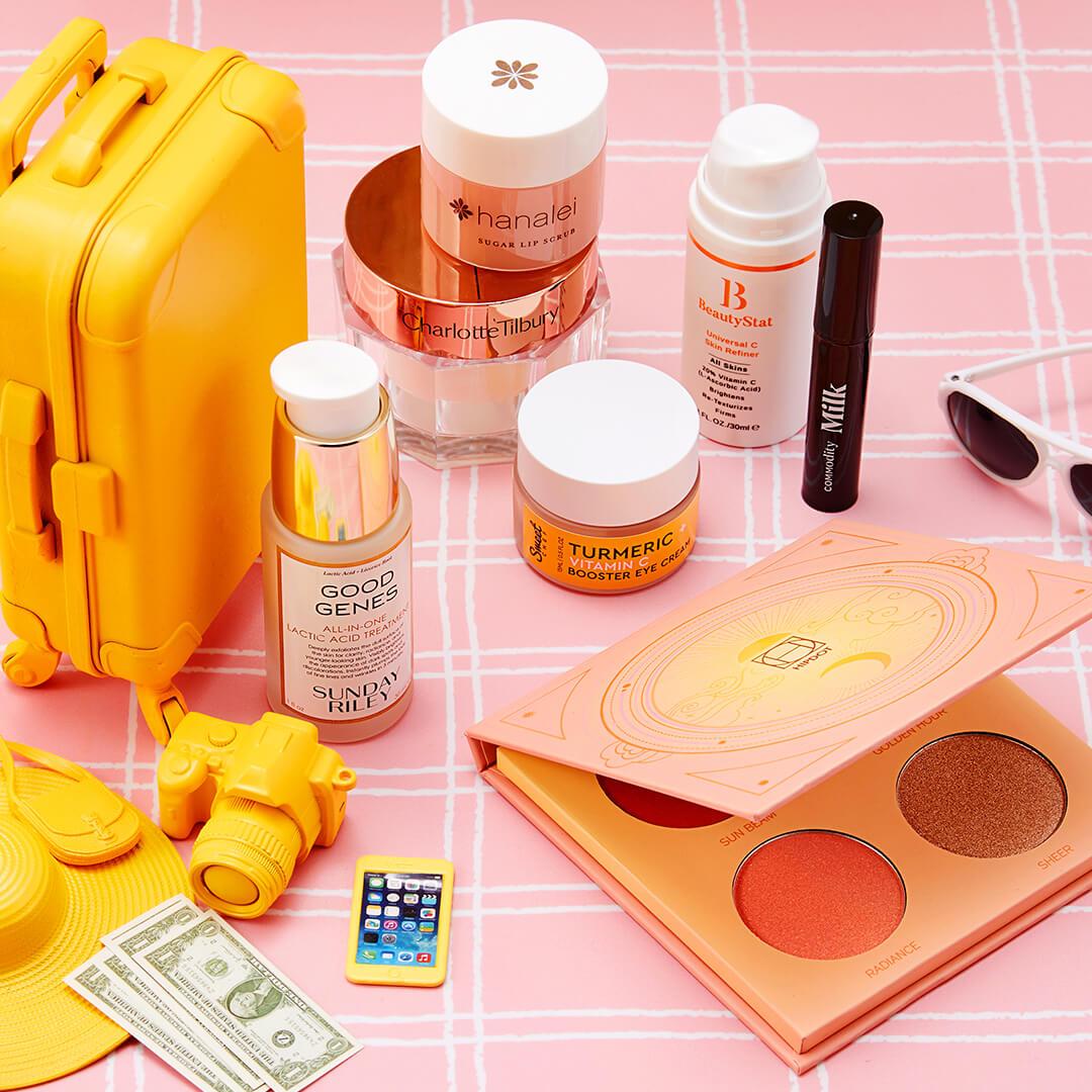 Flat-lay image of makeup and skincare products from various brands, miniature yellow luggage, camera, hat, dollar bills, and iPhone on pink and white tile background Mobile