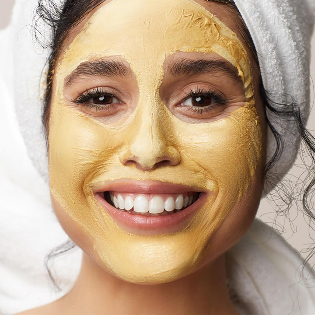 Close-up of a smiling model with her hair wrapped in a towel and yellow face mask on her face Mobile