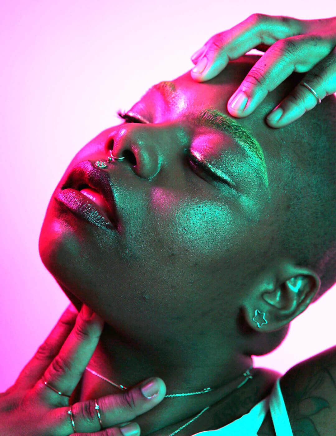 A close-up photo of a woman with a shaved head, her head slightly tilted upward, set against a background illuminated in a soft pink hue infused with AI-driven skincare Mobile