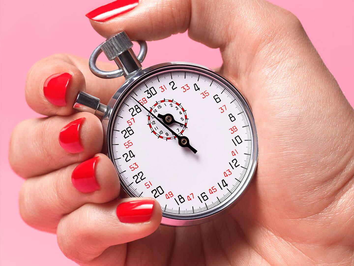 A hand with bold red nail polish holds a silver stopwatch, set against a bright pink background Desktop
