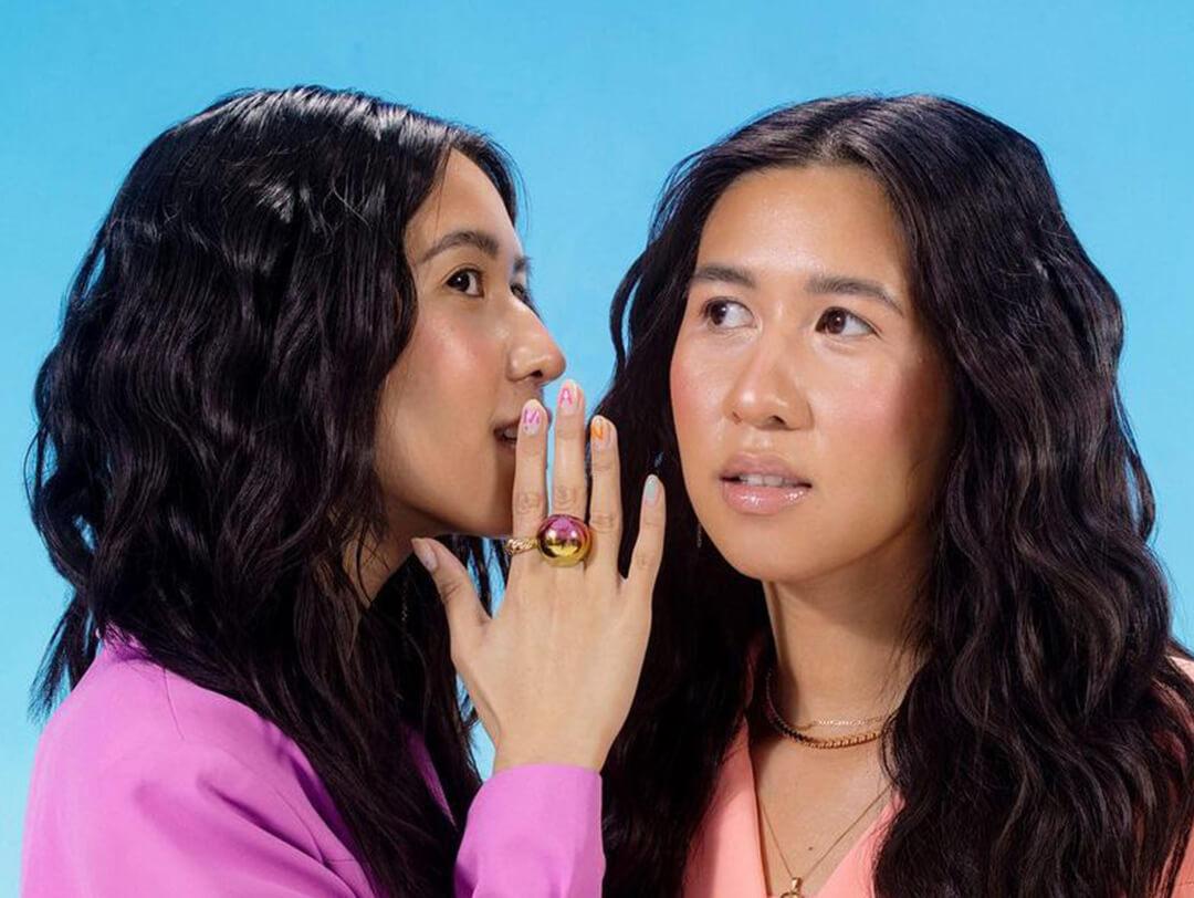 A photo of two models with black wavy hair gossiping to each other on a blue background Desktop