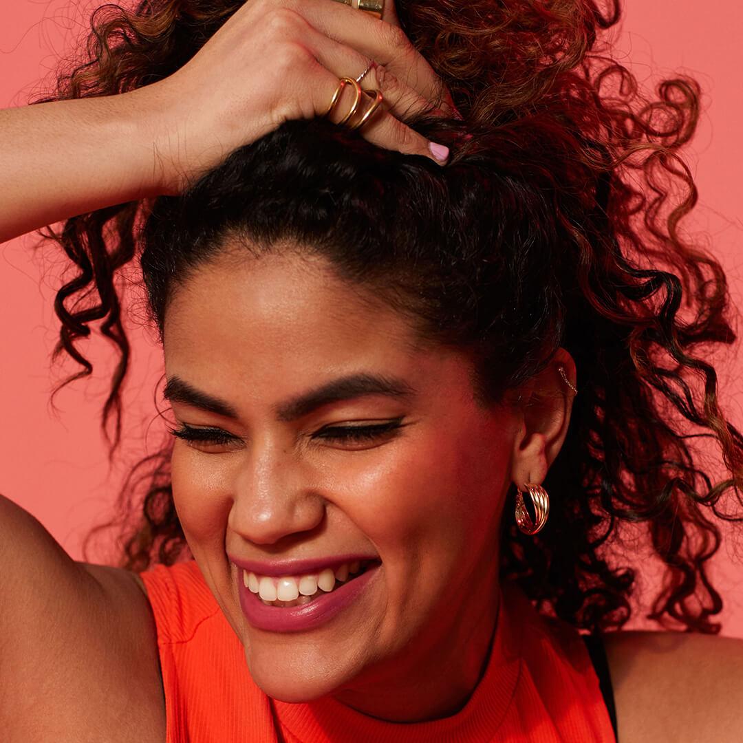 Close-up image of a model with thick, curly hair rocking a natural makeup look wearing an orange top on a red background Desktop