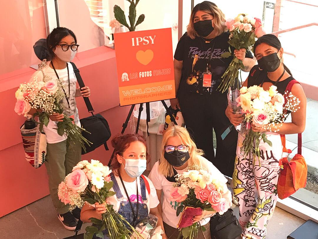 Photo of a group of female photographers with their face masks on each holding a bouquet of flowers around a sign that says "IPSY Las Fotos Project Welcome Photographers" Desktop