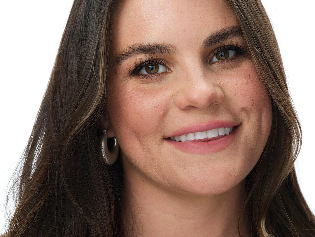 A close-up photo of a woman smiling and looking into the camera, wearing a silver hoop earring and a touch of makeup Desktop